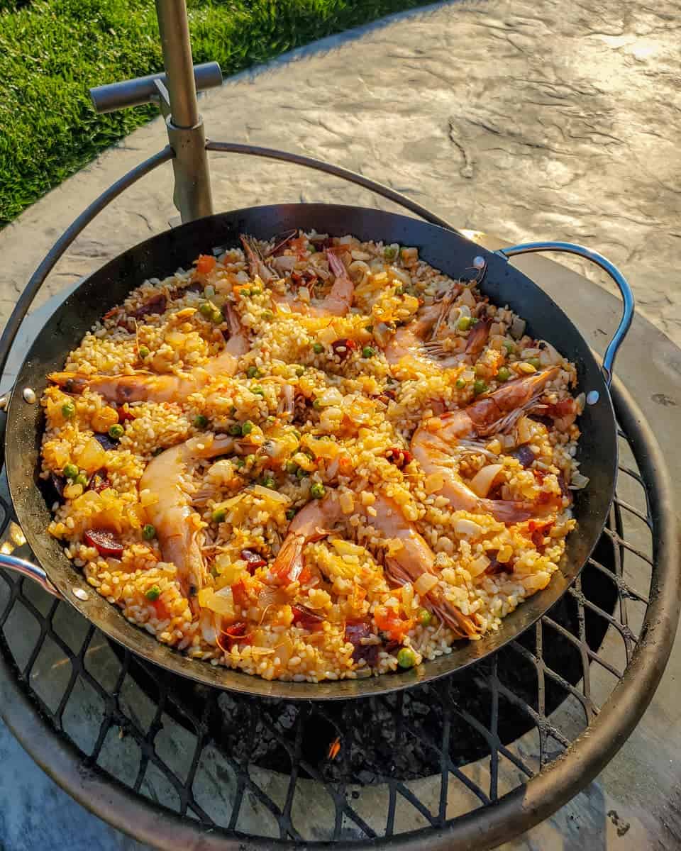 A large skillet of Spanish seafood paella with shrimp, resting on a circular outdoor grill, illuminated by sunset light.