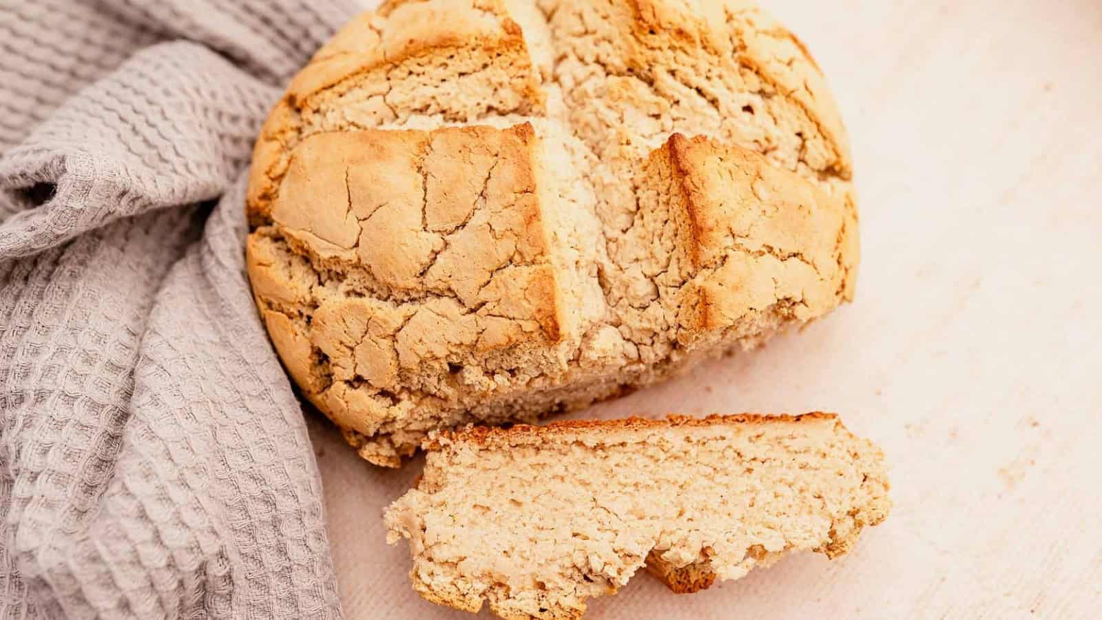 Round loaf of rustic bread with a slice cut off, resting on a light-colored surface next to a gray, textured cloth.