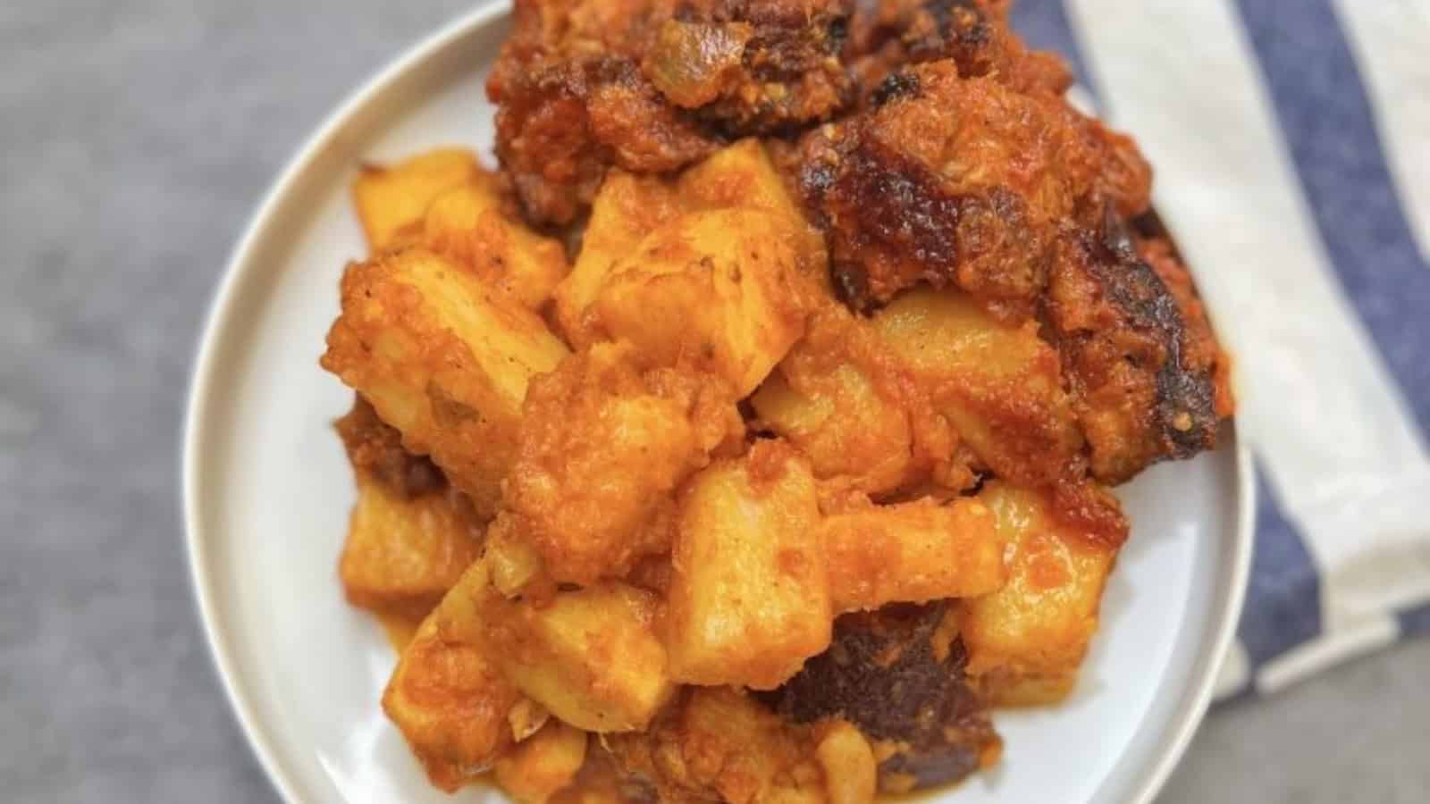 A white plate with fried plantains and spicy meat pieces, placed on a surface with a striped cloth nearby.