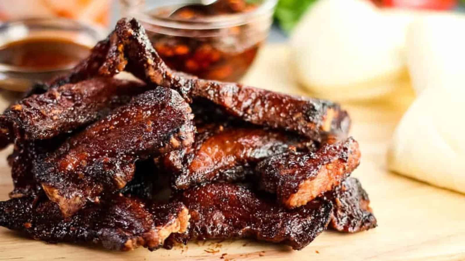 A stack of grilled beef ribs on a wooden board, with a jar of sauce and two steamed buns in the background.