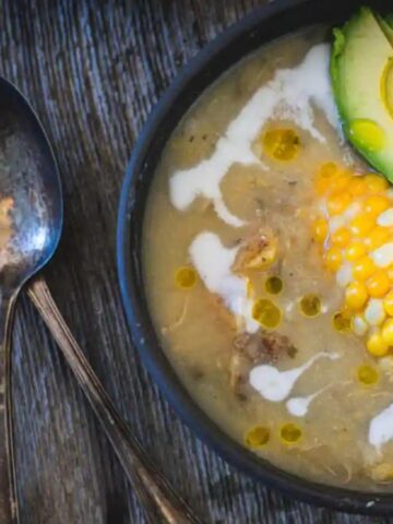 A bowl of creamy soup topped with corn, avocado slices, and a drizzle of oil, beside two spoons on a wooden surface.