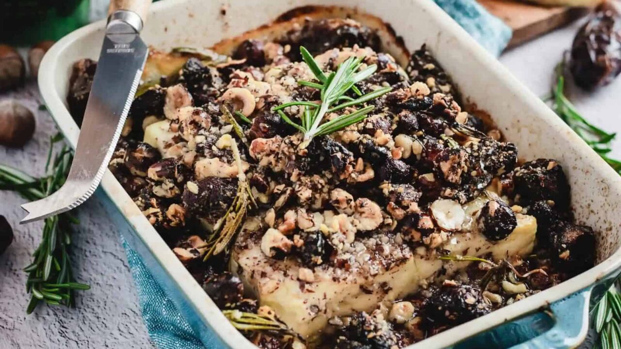 A baked dish with soft cheese topped with chopped nuts, dried fruits, and a sprig of rosemary in a rectangular white baking dish.