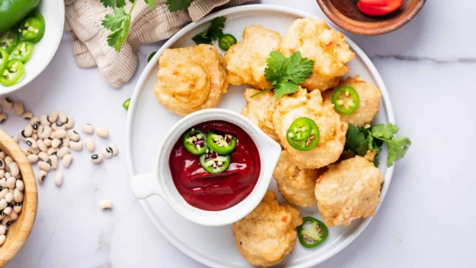 A white plate with fried fritters garnished with cilantro and sliced jalapeños alongside a bowl of red sauce topped with jalapeño slices, placed on a marble surface.