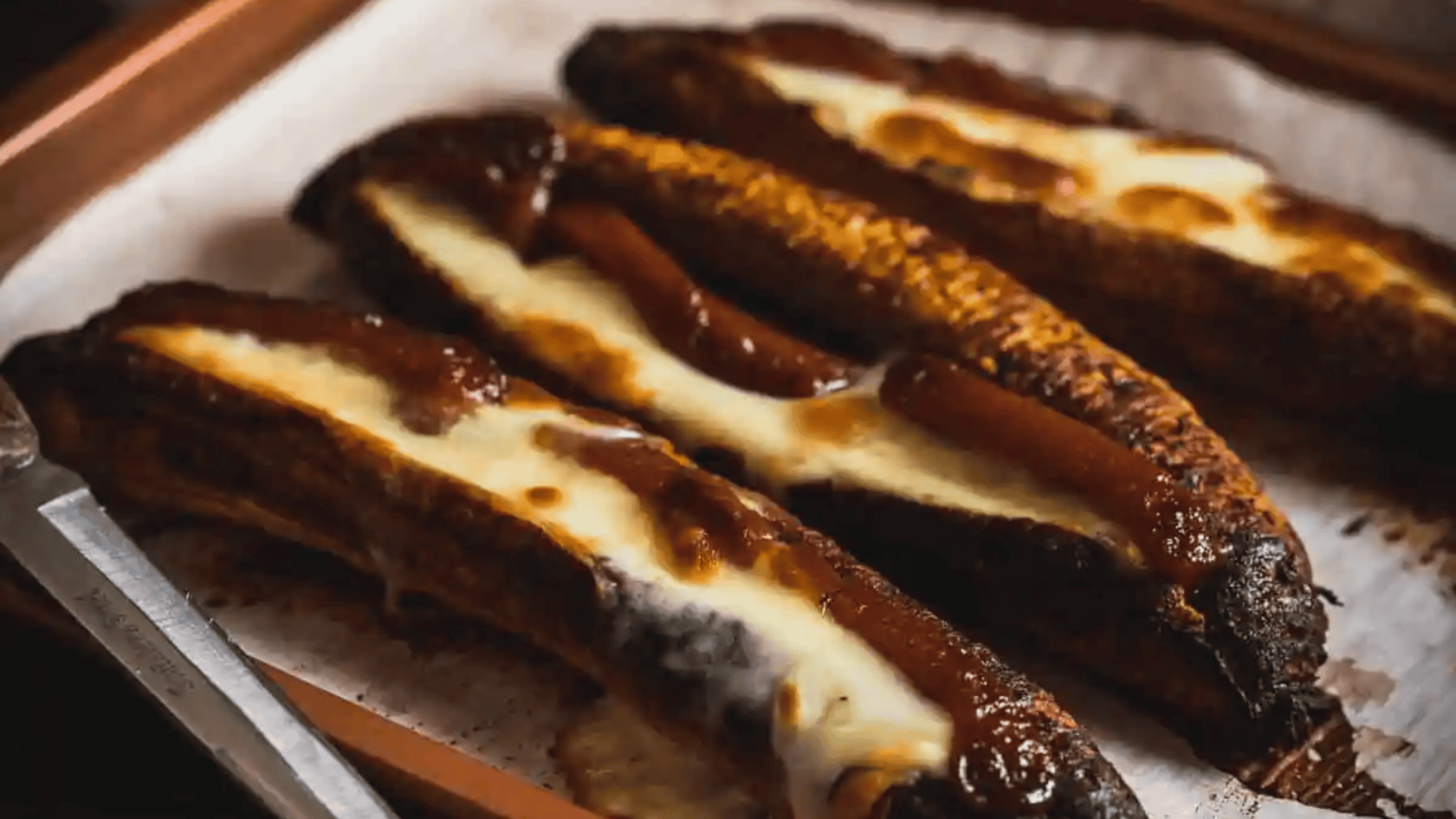 Three baked bananas topped with melted chocolate and cheese on a parchment-lined tray.