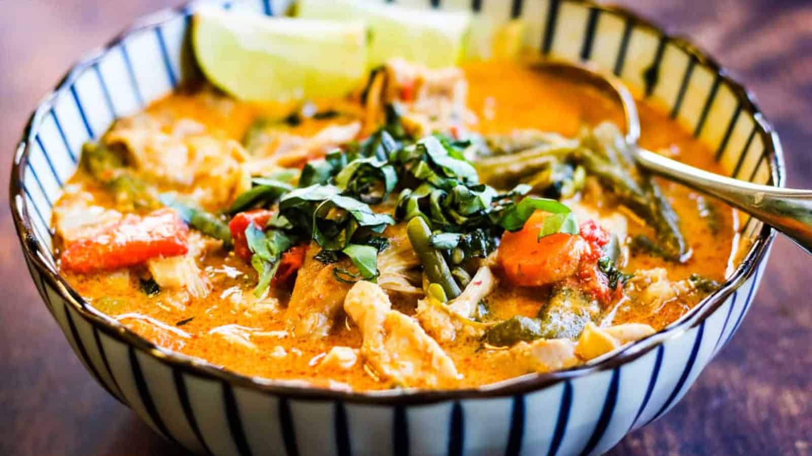 A bowl of Thai red curry with chicken, bell peppers, and basil, garnished with lime wedges. A spoon rests in the bowl.