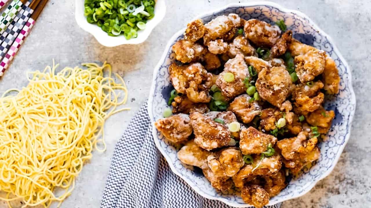 A bowl of crispy fried chicken garnished with green onions is on a table next to a small pile of uncooked noodles and a bowl of chopped green onions. A striped napkin is partially visible.