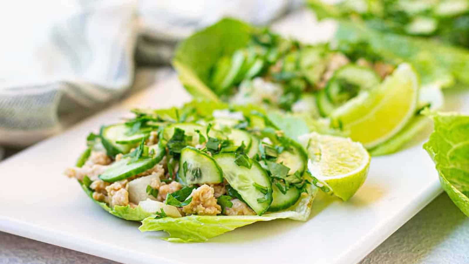 Lettuce wraps filled with minced meat, sliced cucumbers, and chopped herbs on a white plate, garnished with lime wedges.