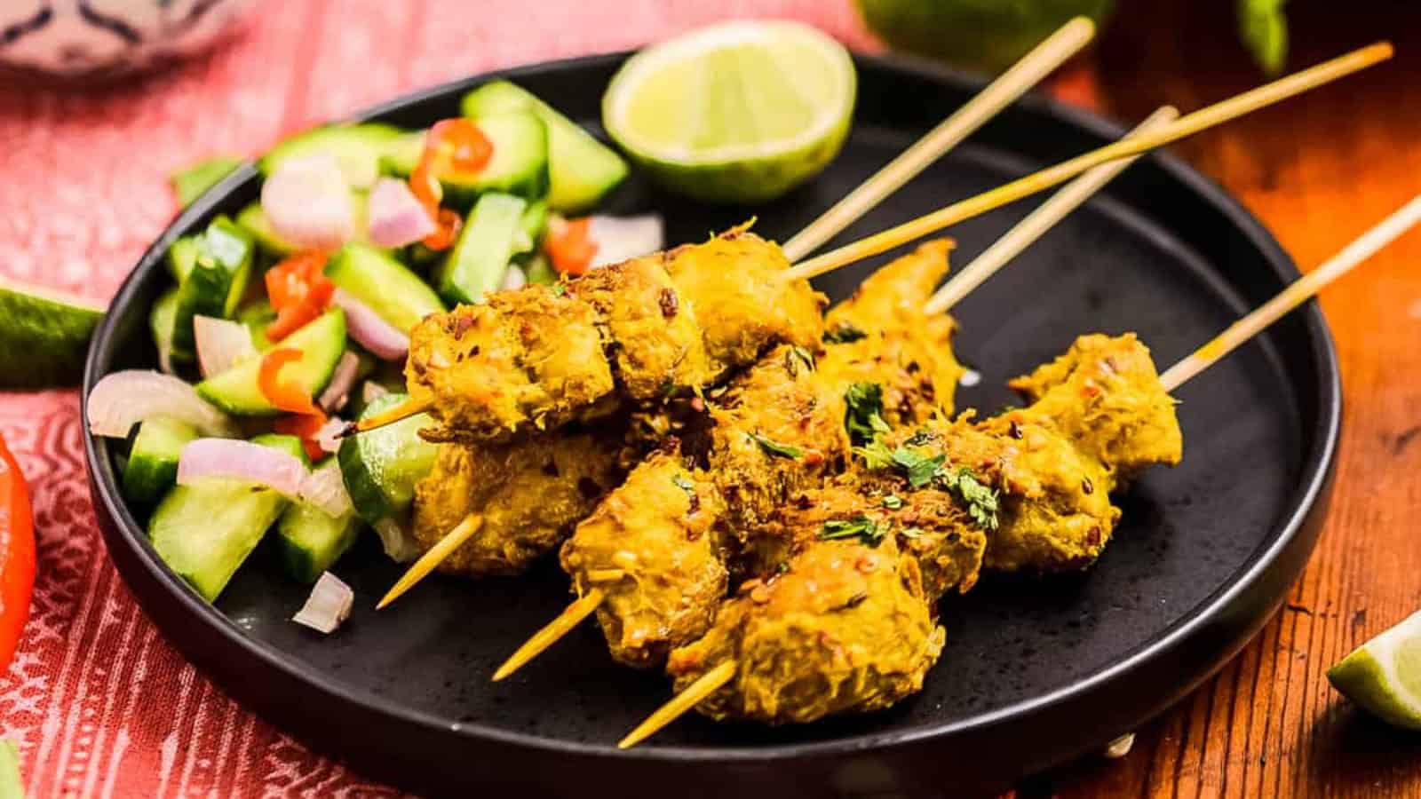 Chicken skewers with herbs on a black plate, served with a side of cucumber and onion salad, and a lime wedge.