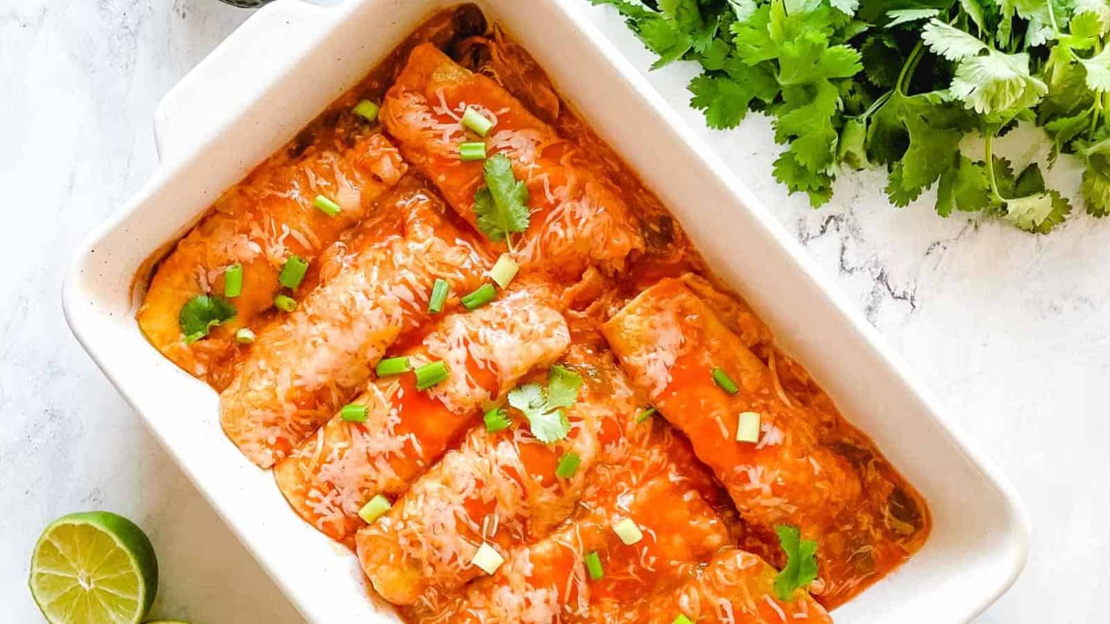A baking dish full of chicken enchiladas is surrounded by cut limes, a halved avocado, and a green linen.