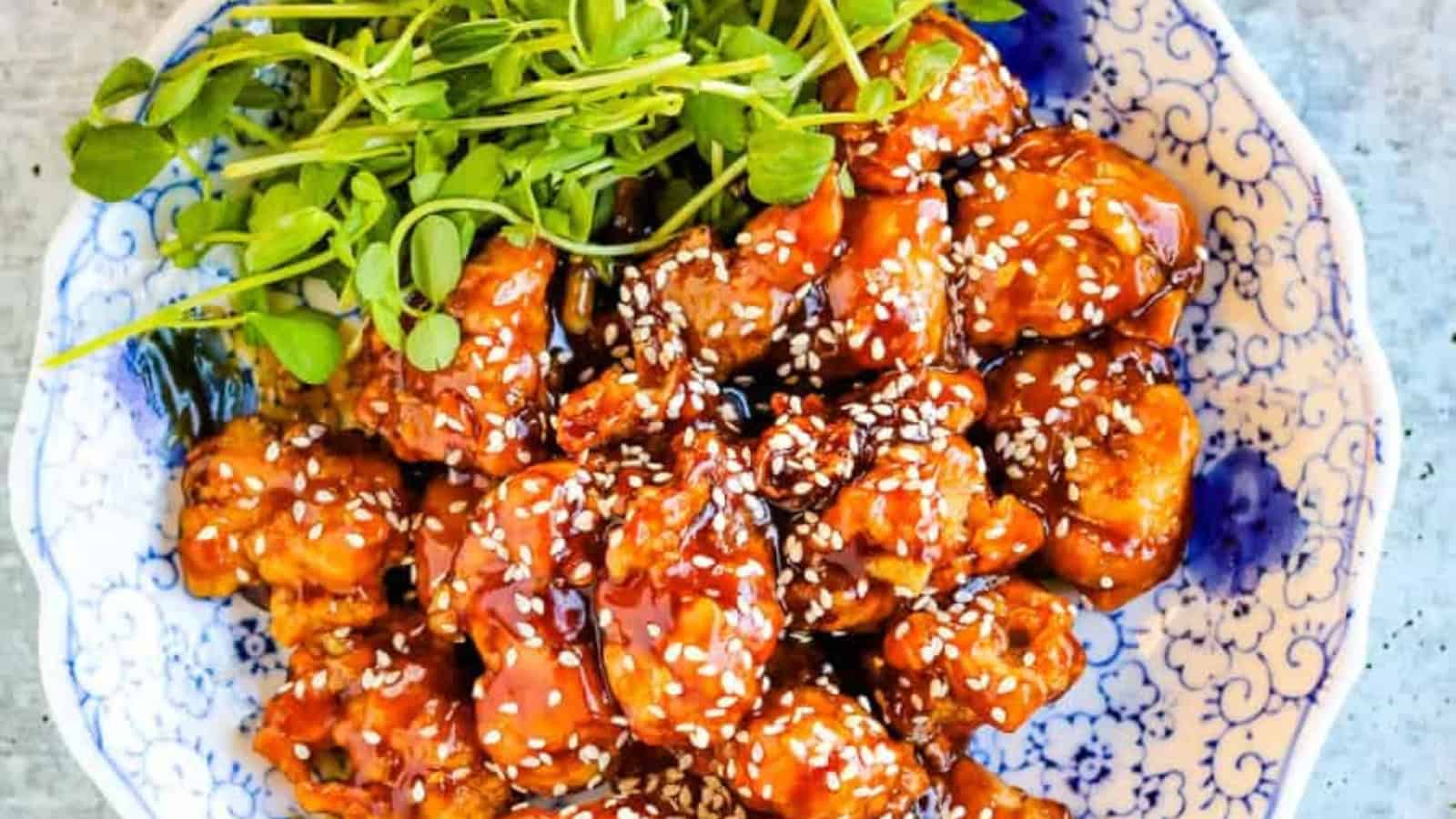 Plate of glazed sesame chicken topped with white sesame seeds, accompanied by fresh greens, served on a decorative blue and white dish.