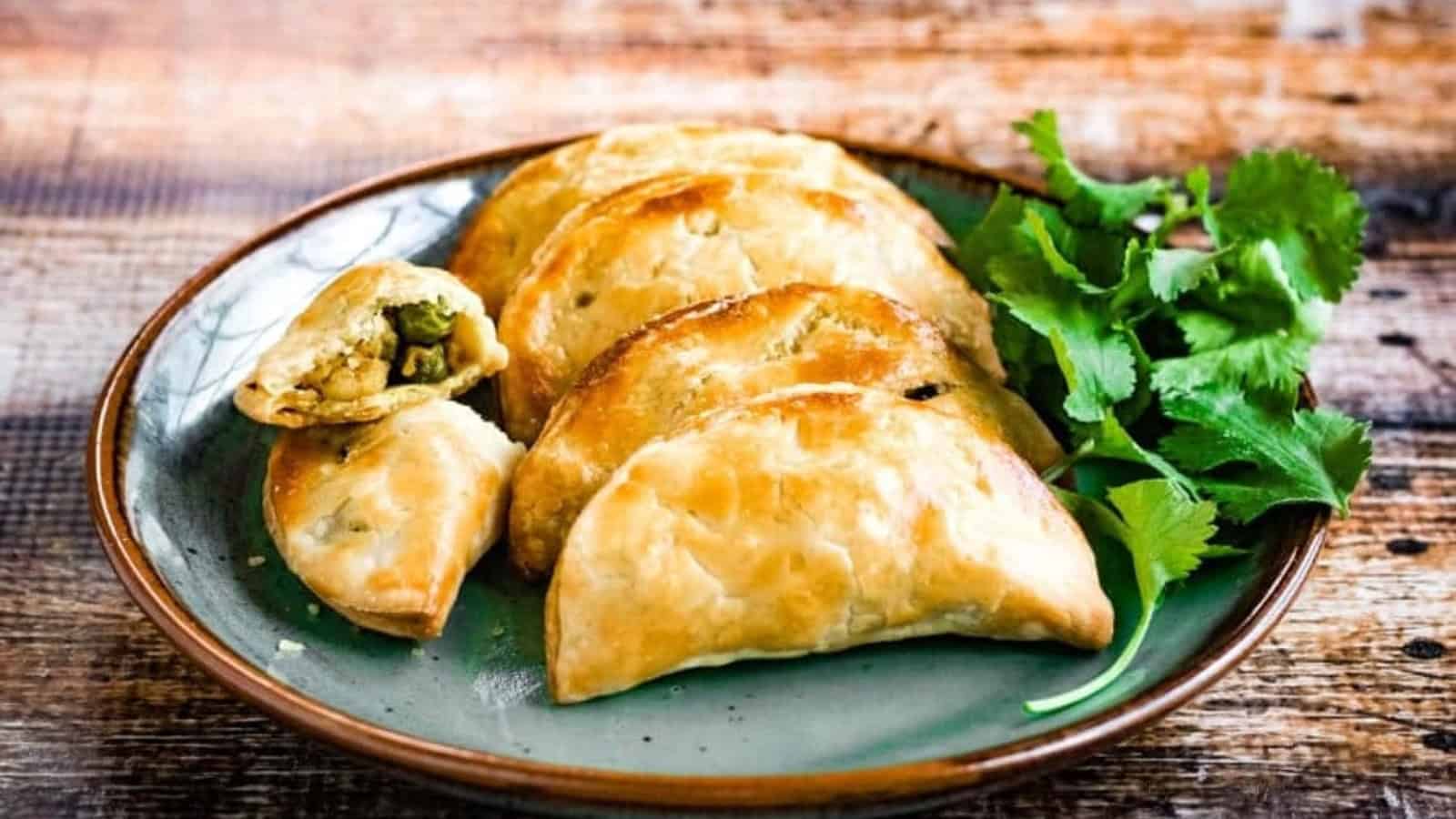 A plate of baked empanadas with a side of fresh cilantro. One empanada is cut open, showing a vegetable filling.