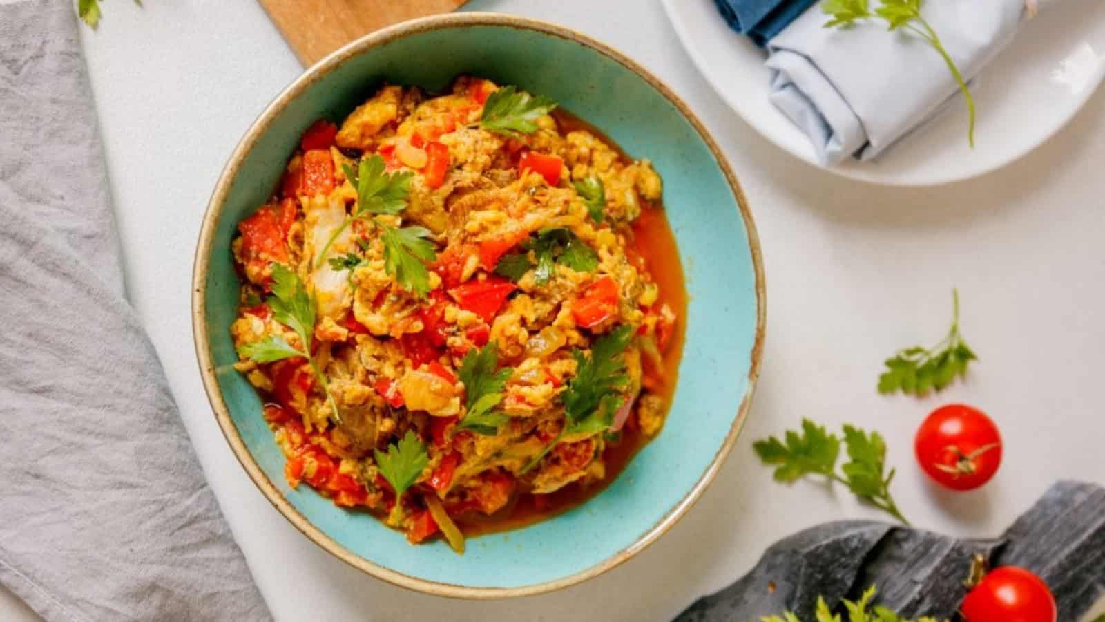 A bowl of vegetable stir-fry with tomatoes and herbs is on a table, accompanied by a cloth napkin and fresh cherry tomatoes.