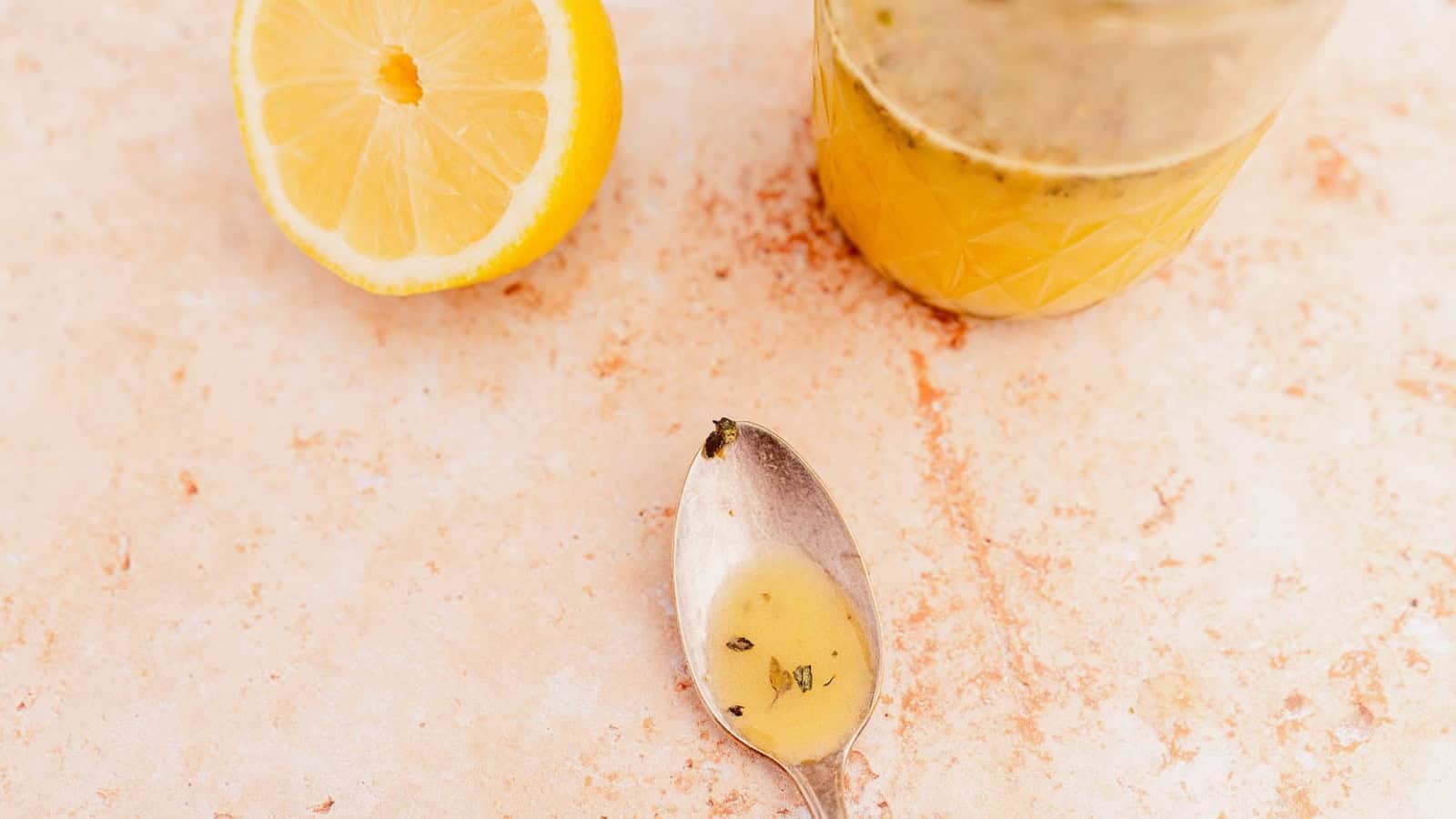 A spoon with dressing next to a sliced lemon and a jar on a textured surface.