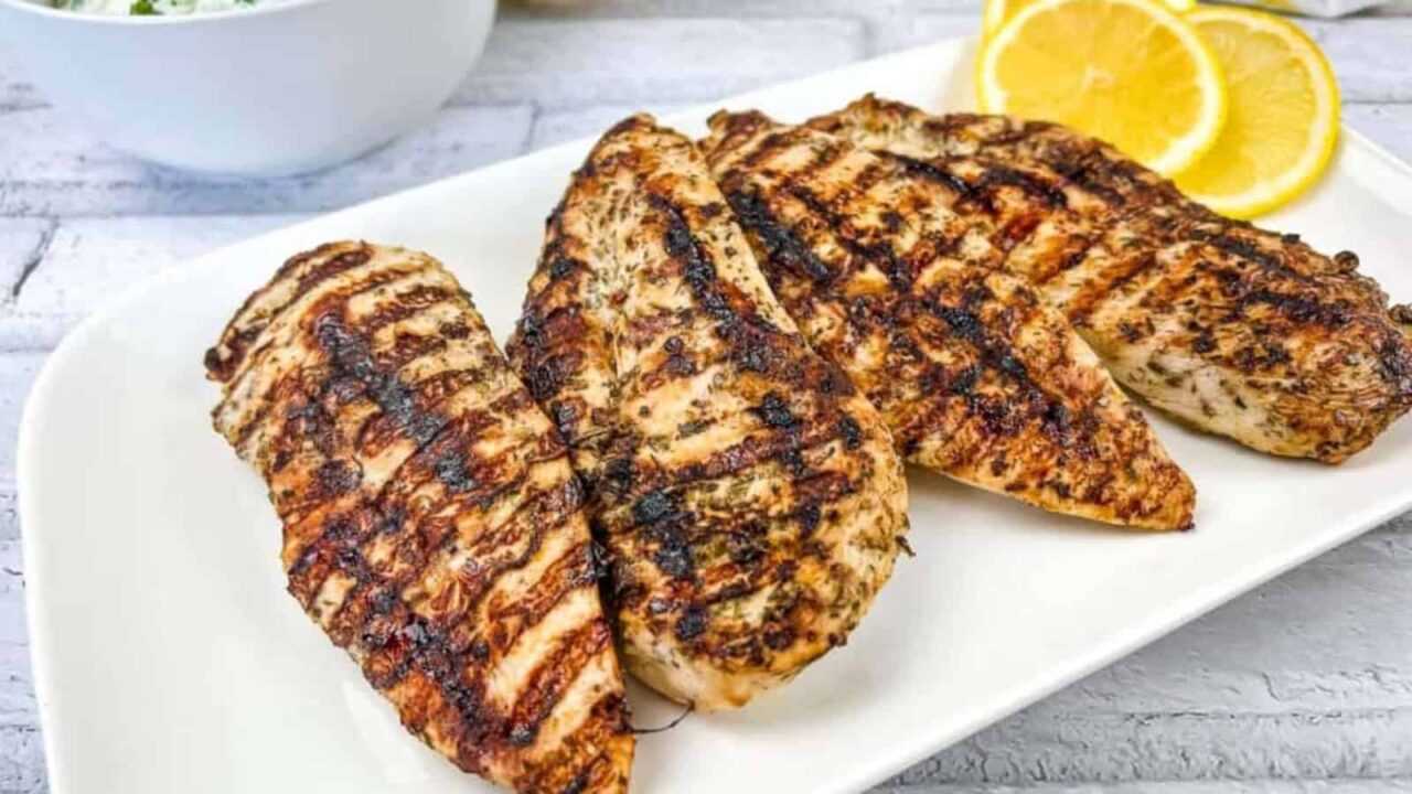 Grilled chicken breasts on a white plate with lemon slices on the side.