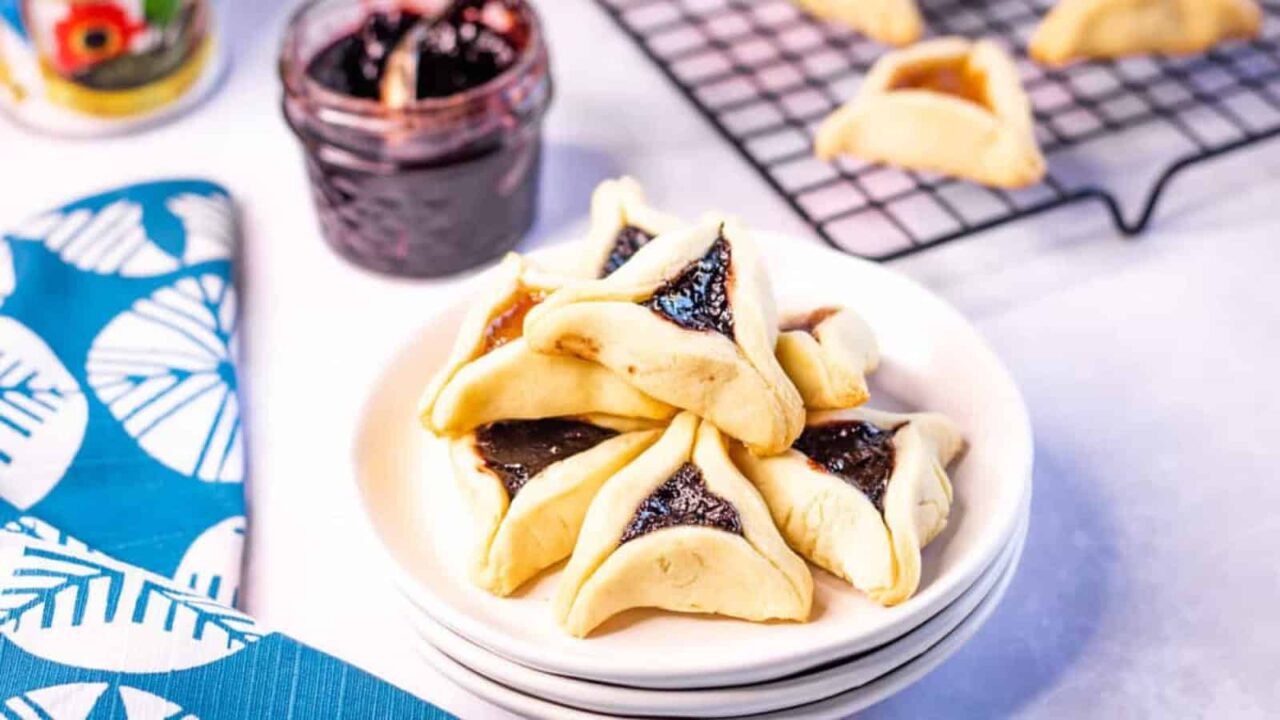 Hamentashen cookies piled on a white plate with more cookies on a rack in the background. There is jar of jam with a spoon in it in the background too.