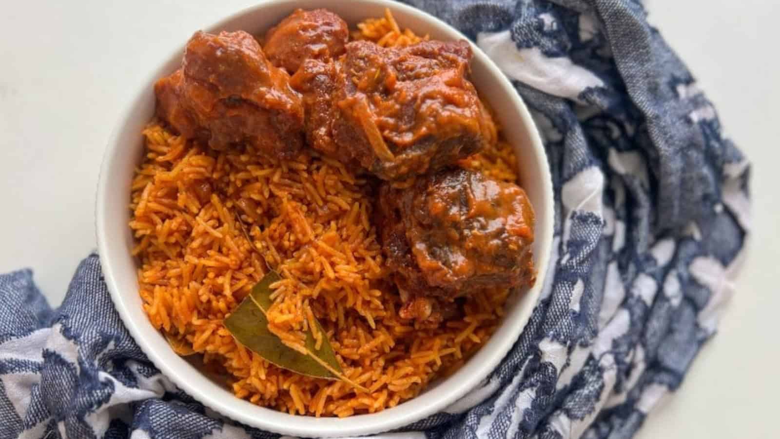 A bowl of jollof rice topped with meat pieces, garnished with a bay leaf, placed on a patterned cloth.
