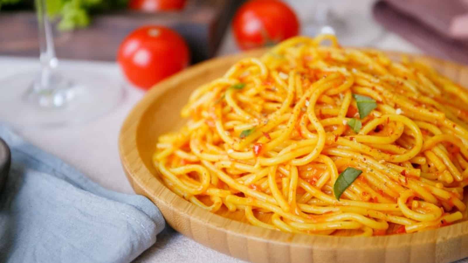 A wooden plate of spaghetti with tomato sauce, garnished with basil and black pepper, is on a table with whole tomatoes and a glass of wine visible in the background.