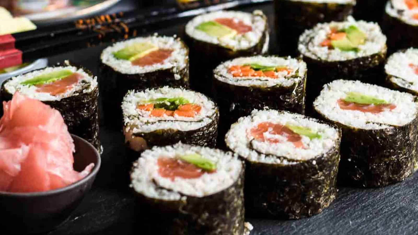 Close-up of sushi rolls with various fillings, including avocado and salmon, arranged on a black surface with a side of pink pickled ginger.