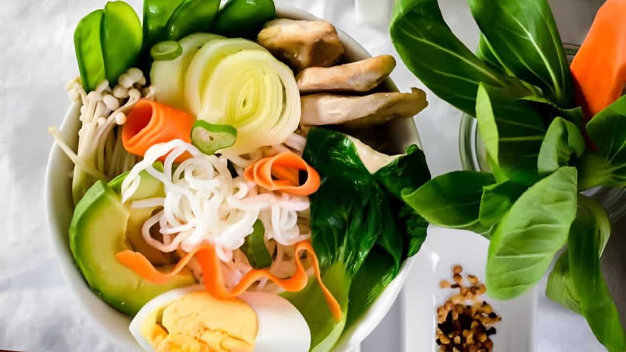 A bowl of ramen with sliced avocado, boiled egg, mushrooms, bok choy, carrots, snow peas, and noodles on a white surface.