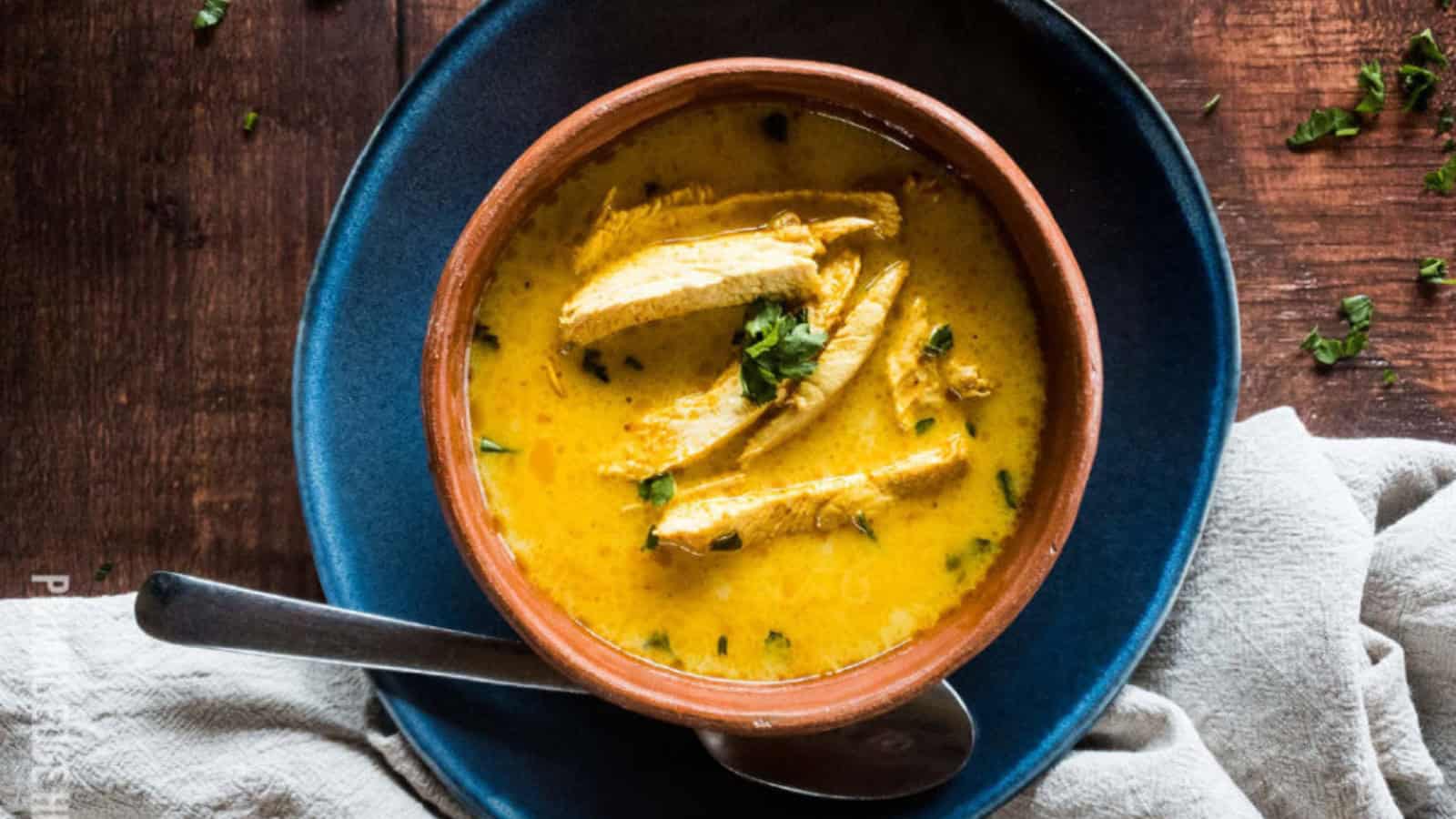 Bowl of creamy chicken curry with cilantro garnish on a wooden table, accompanied by a silver spoon and cloth napkin.