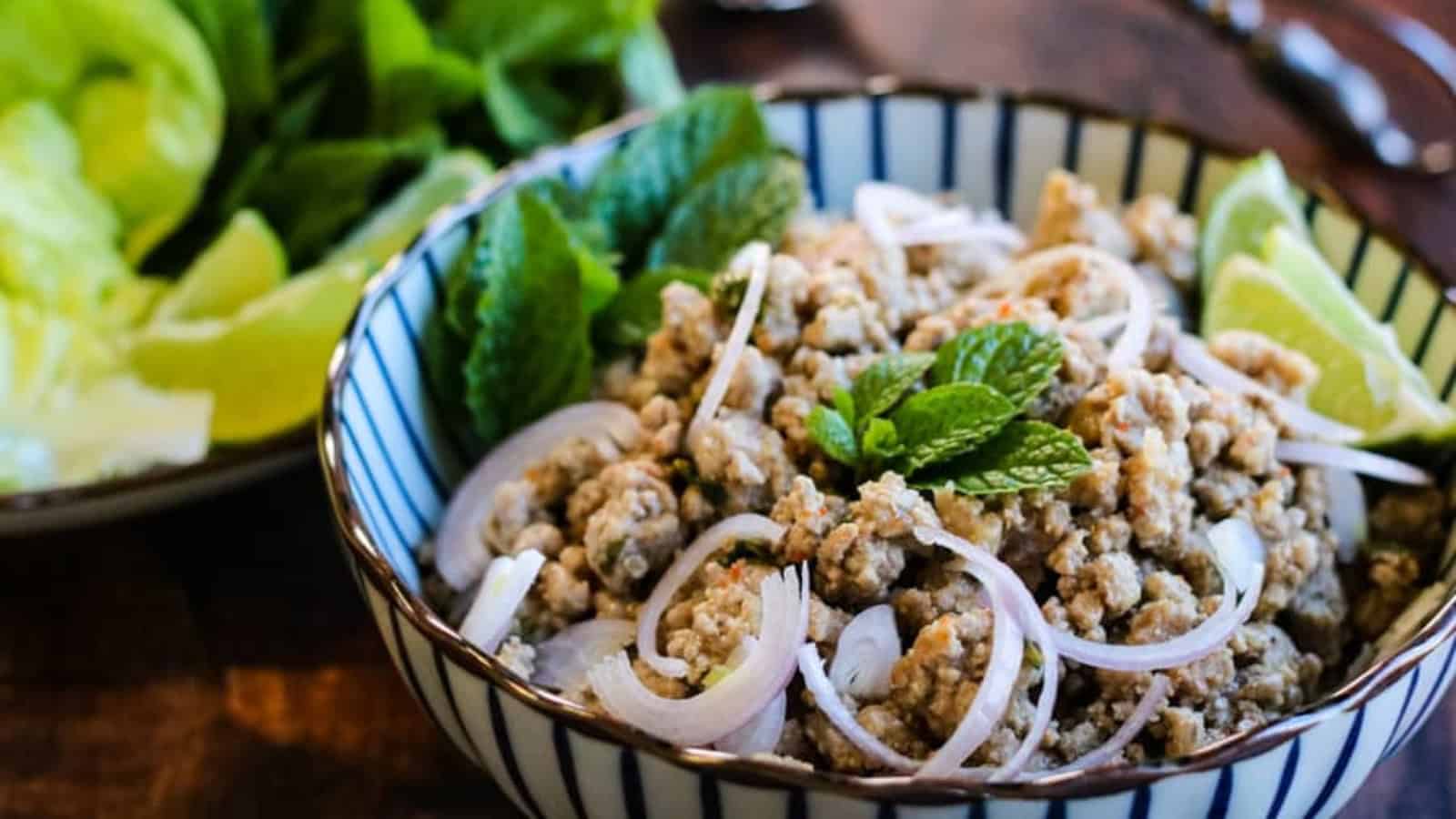 A bowl of seasoned ground meat topped with sliced onions and fresh mint leaves, accompanied by lime wedges. Leaves are visible in the background.