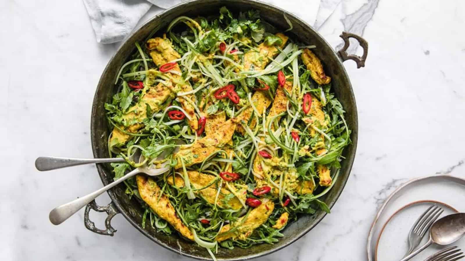 A large pan filled with a salad featuring sliced chicken, leafy greens, avocado, red chili slices, and thinly sliced vegetables, with two serving spoons on a white marble surface.