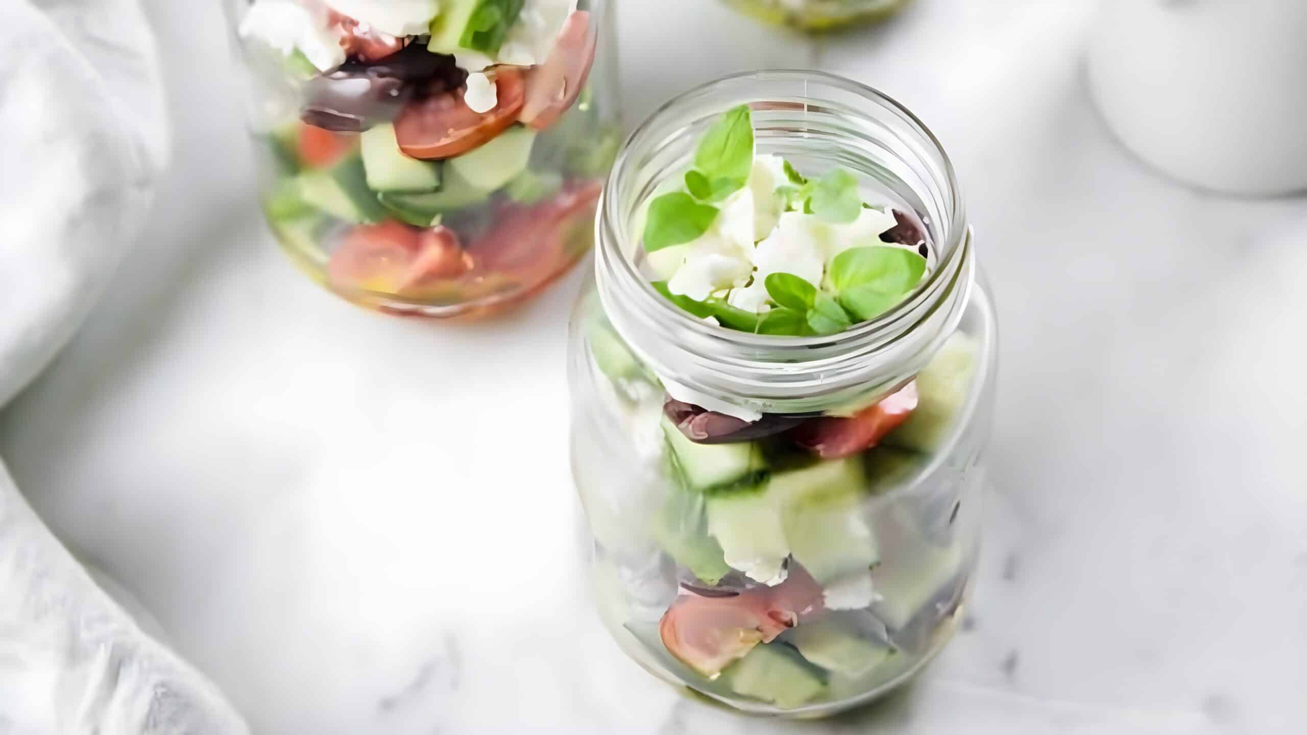 Two glass jars filled with salad, containing cucumbers, tomatoes, olives, feta cheese, and basil leaves, placed on a marble surface.