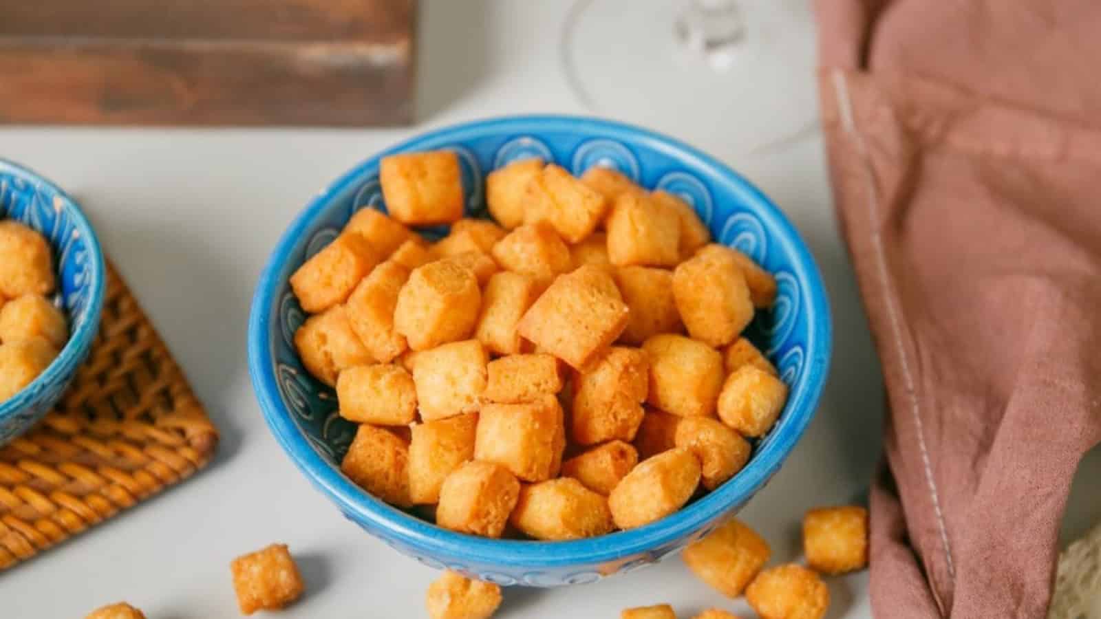 A blue bowl filled with golden brown, cube-shaped snacks on a table, with a cloth napkin beside it.
