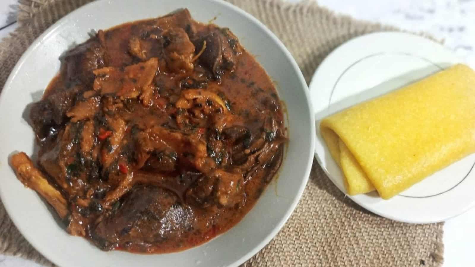 A plate of spicy stew with assorted meats next to a plate of yellow cornmeal porridge.