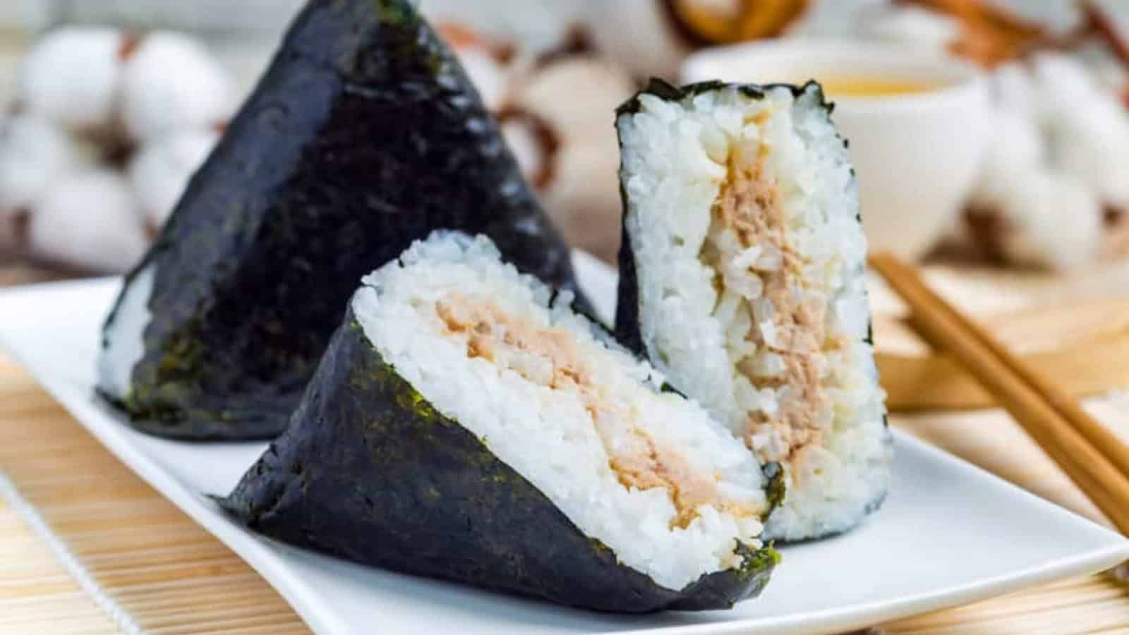 Three rice balls wrapped in seaweed, filled with tuna, on a white plate with chopsticks nearby.