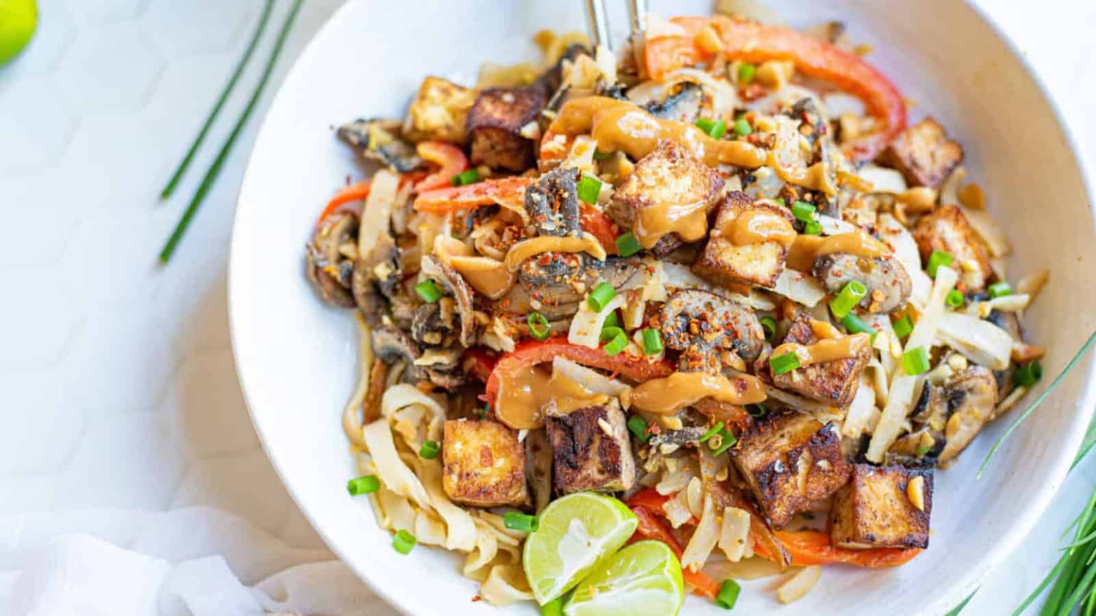 A bowl of tofu stir-fry with vegetables, topped with a creamy sauce, garnished with chopped green onions and lime wedges.