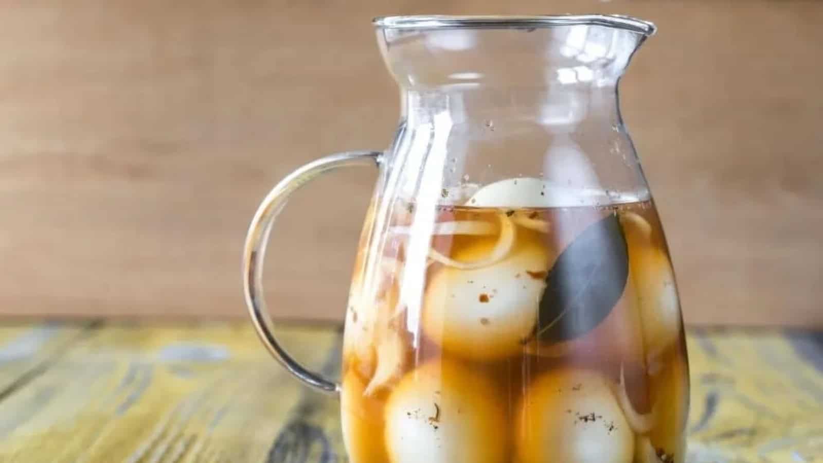 Glass pitcher filled with hard-boiled eggs in brown liquid, containing spices and a bay leaf, placed on a wooden surface.