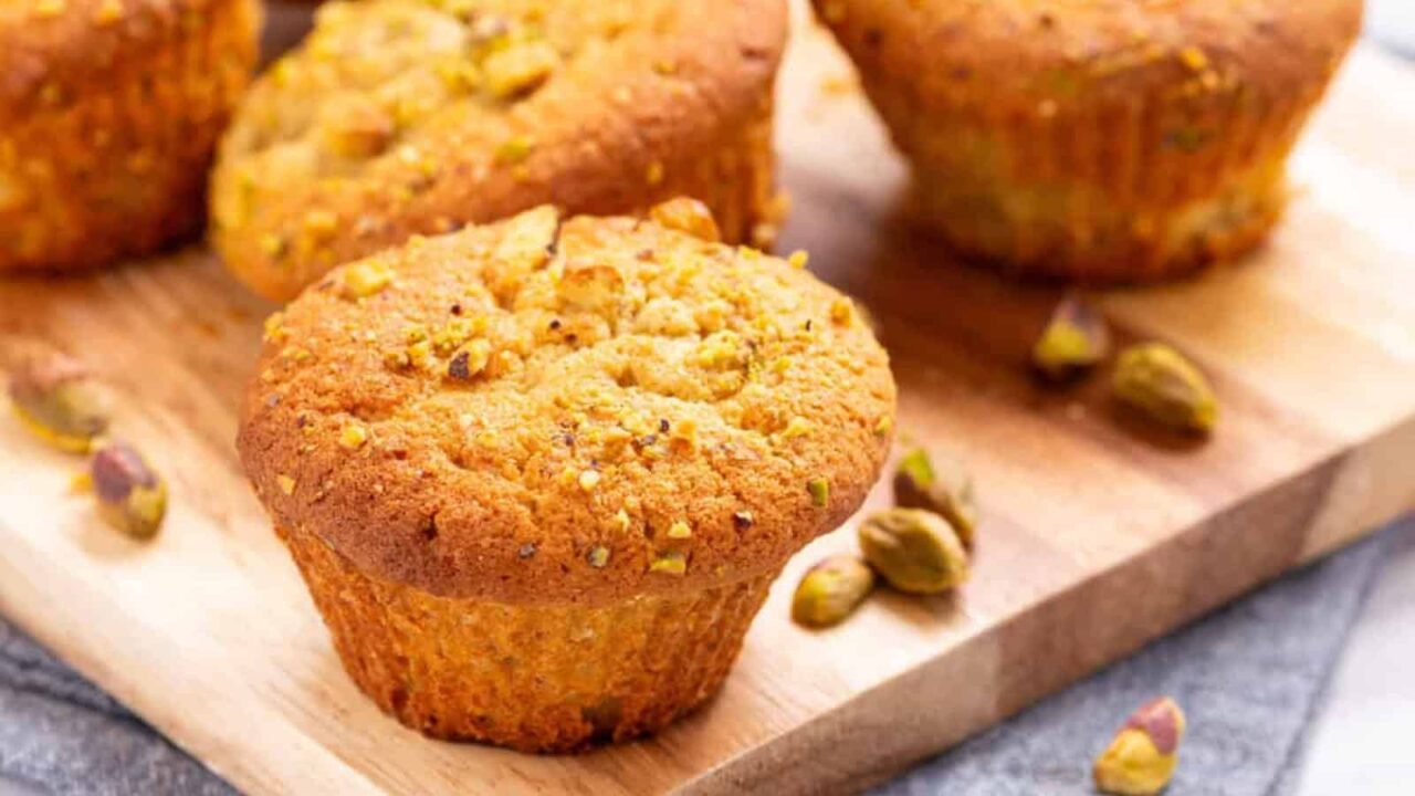 Top view of four pistachio muffins on a wooden board, with one muffin placed separately and pistachios in the background.