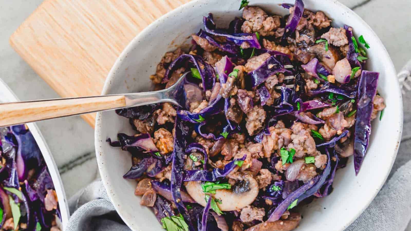 A bowl of cooked ground meat mixed with red cabbage, mushrooms, and herbs, with a wooden-handled spoon resting inside.