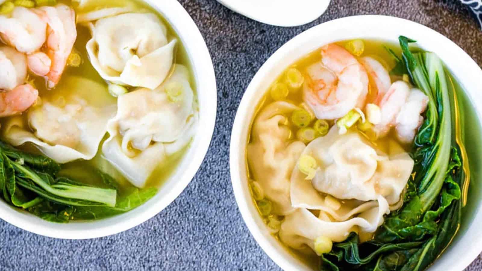 Two bowls of soup with dumplings, shrimp, bok choy, and green onions on a gray surface.