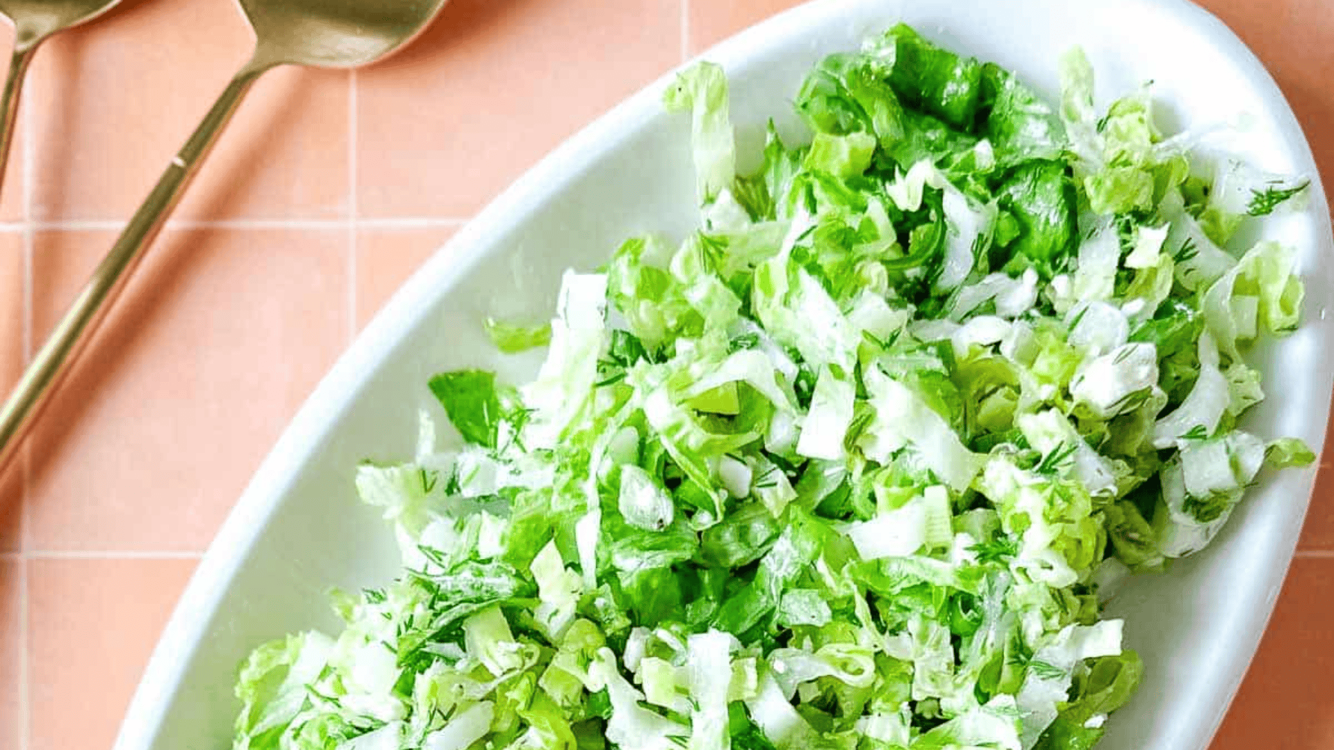 A white oval dish filled with chopped green lettuce and herbs on a tile table. Two gold spoons are placed beside it.