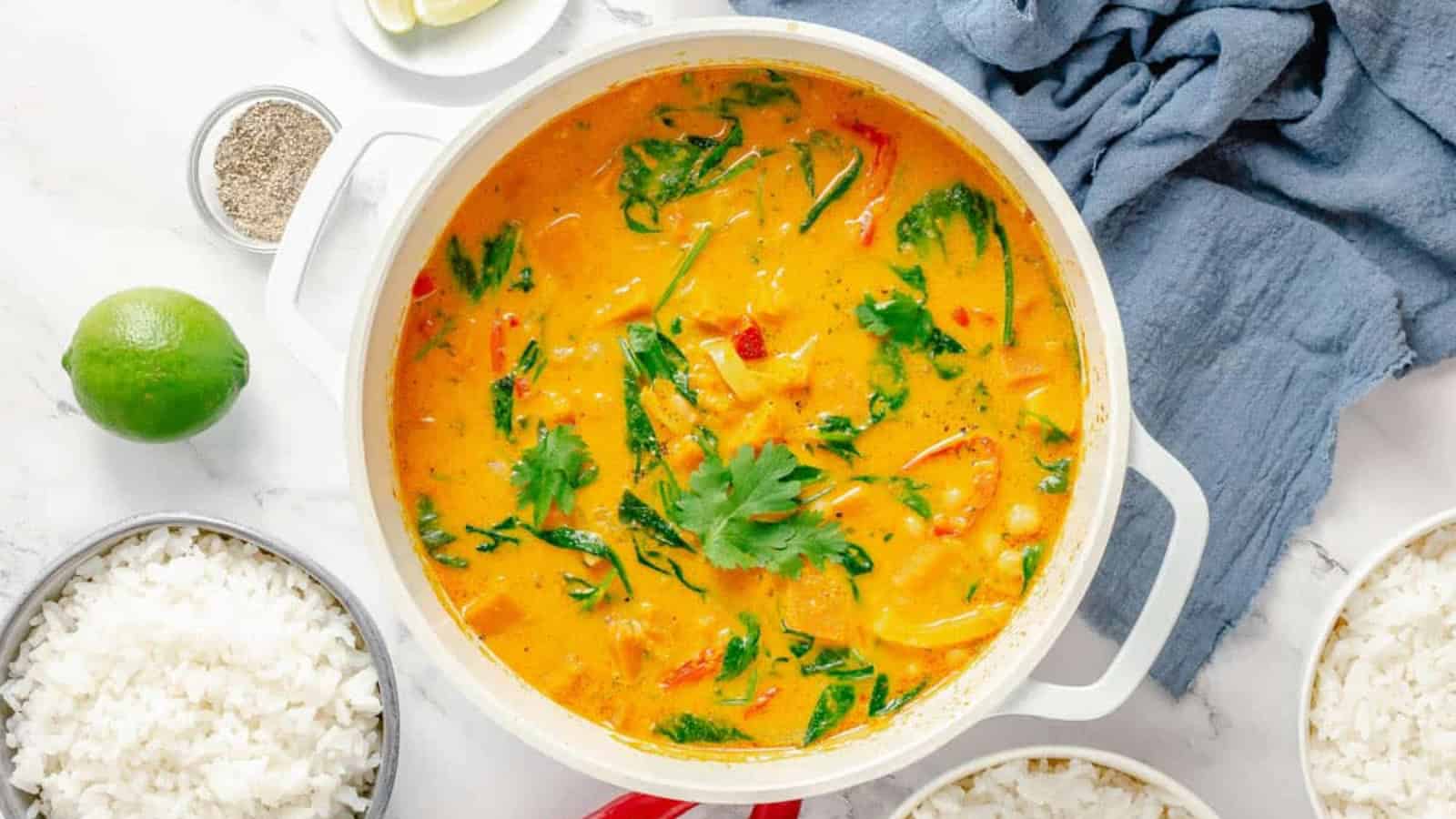 A pot filled with orange curry with vegetables and herbs, surrounded by bowls of white rice, a lime, and a cloth napkin.