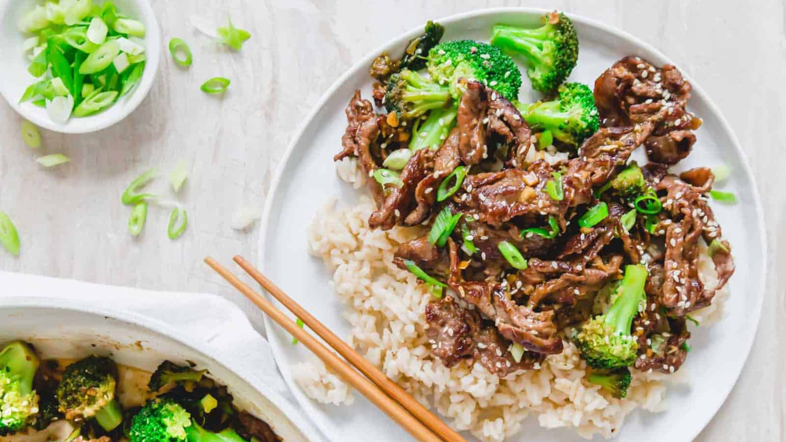 Plate of beef and broccoli with rice, garnished with green onions and chopsticks on the side.