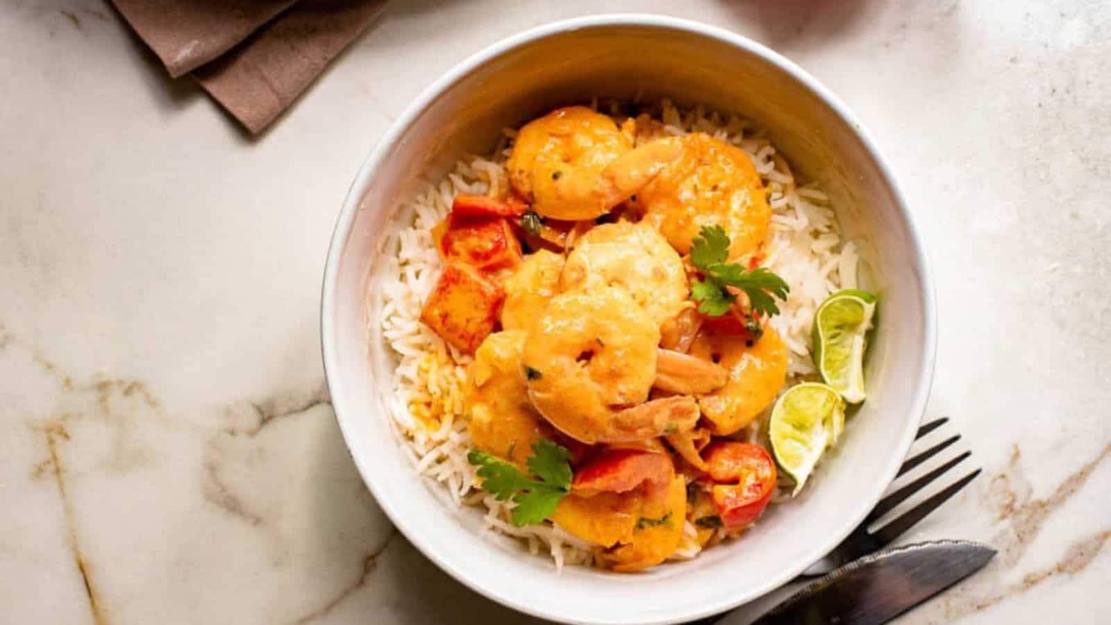A bowl of shrimp curry with rice, garnished with cilantro and lime wedges, placed on a marble surface.