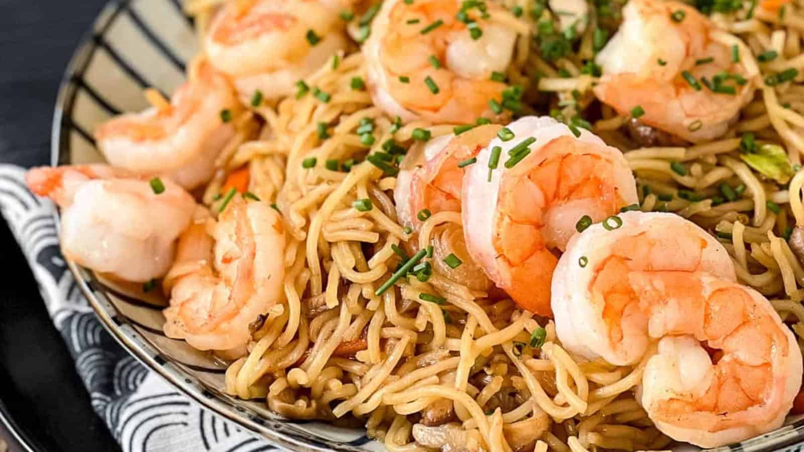 A plate of shrimp lo mein topped with chopped chives on a patterned napkin.