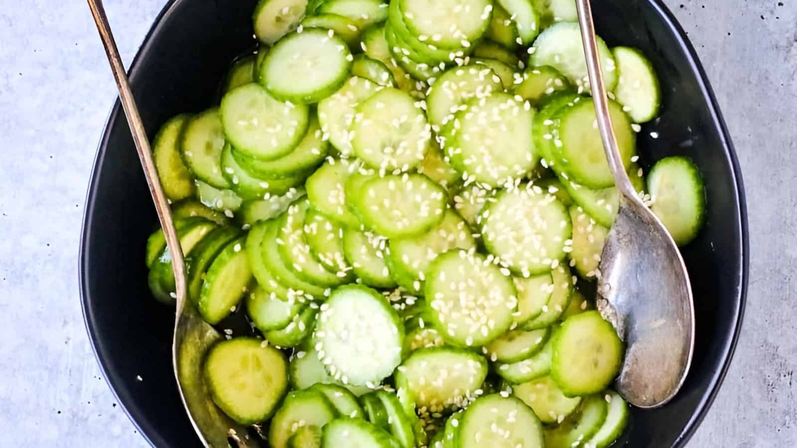 A black bowl filled with sliced cucumbers, sprinkled with sesame seeds, with metal serving utensils on a light surface.
