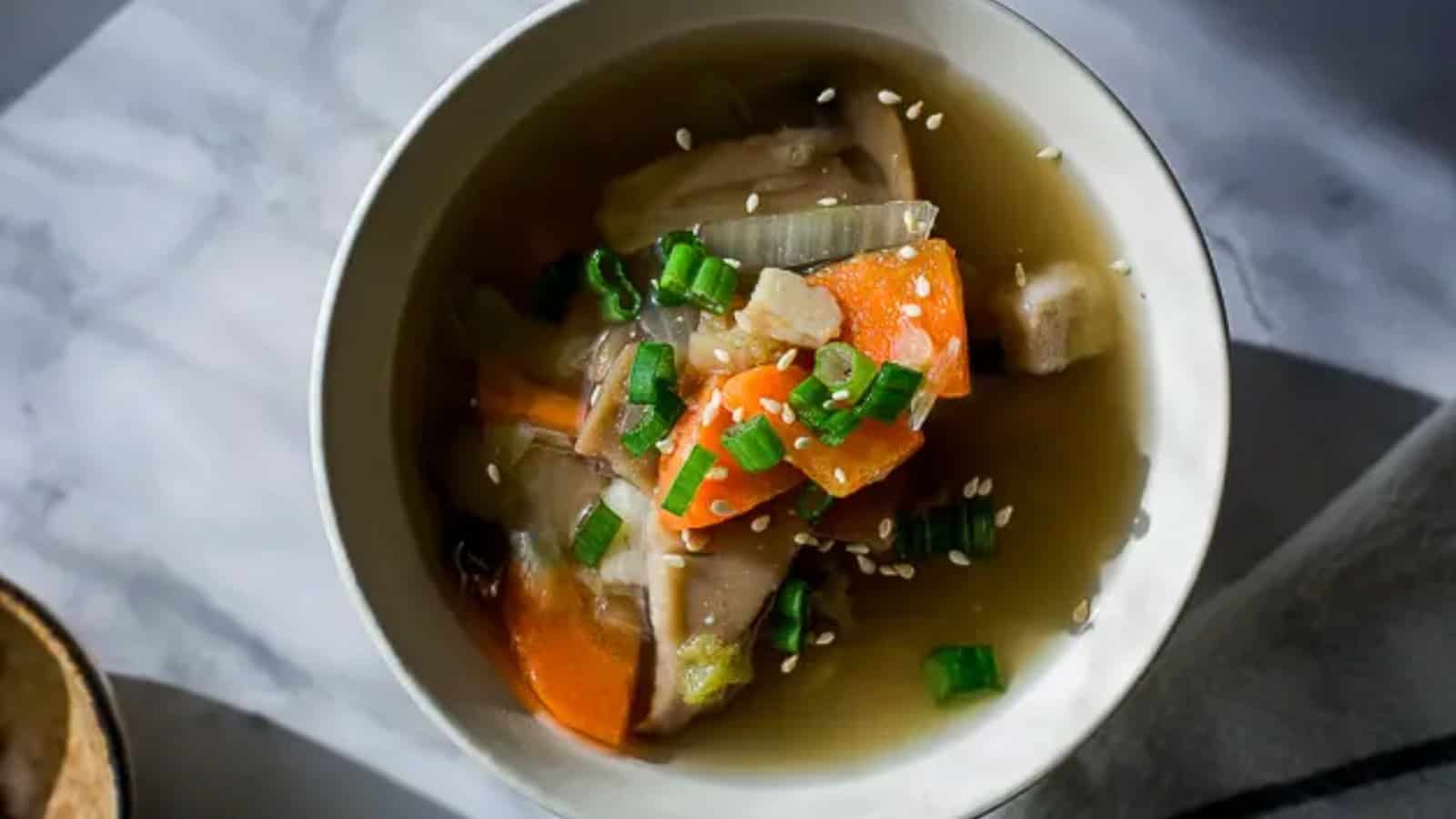 A bowl of miso soup with carrots, mushrooms, onions, green onions, and sesame seeds on a marble surface.