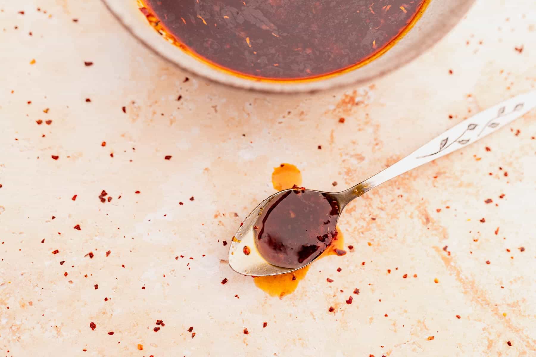 A ceramic bowl and a spoon with dark red sauce on a beige surface, sprinkled with chili flakes.