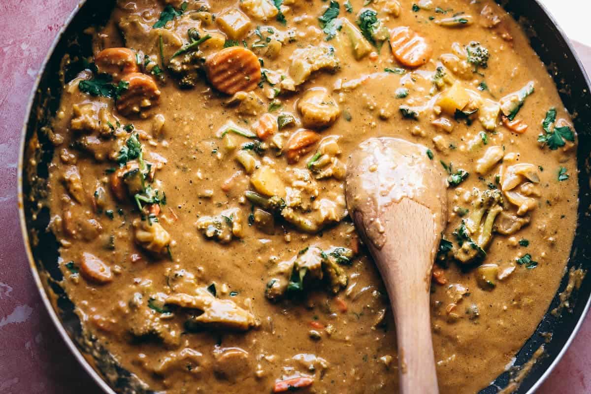 A pan filled with creamy vegetable curry, featuring carrots, broccoli, and herbs, stirred with a wooden spoon.