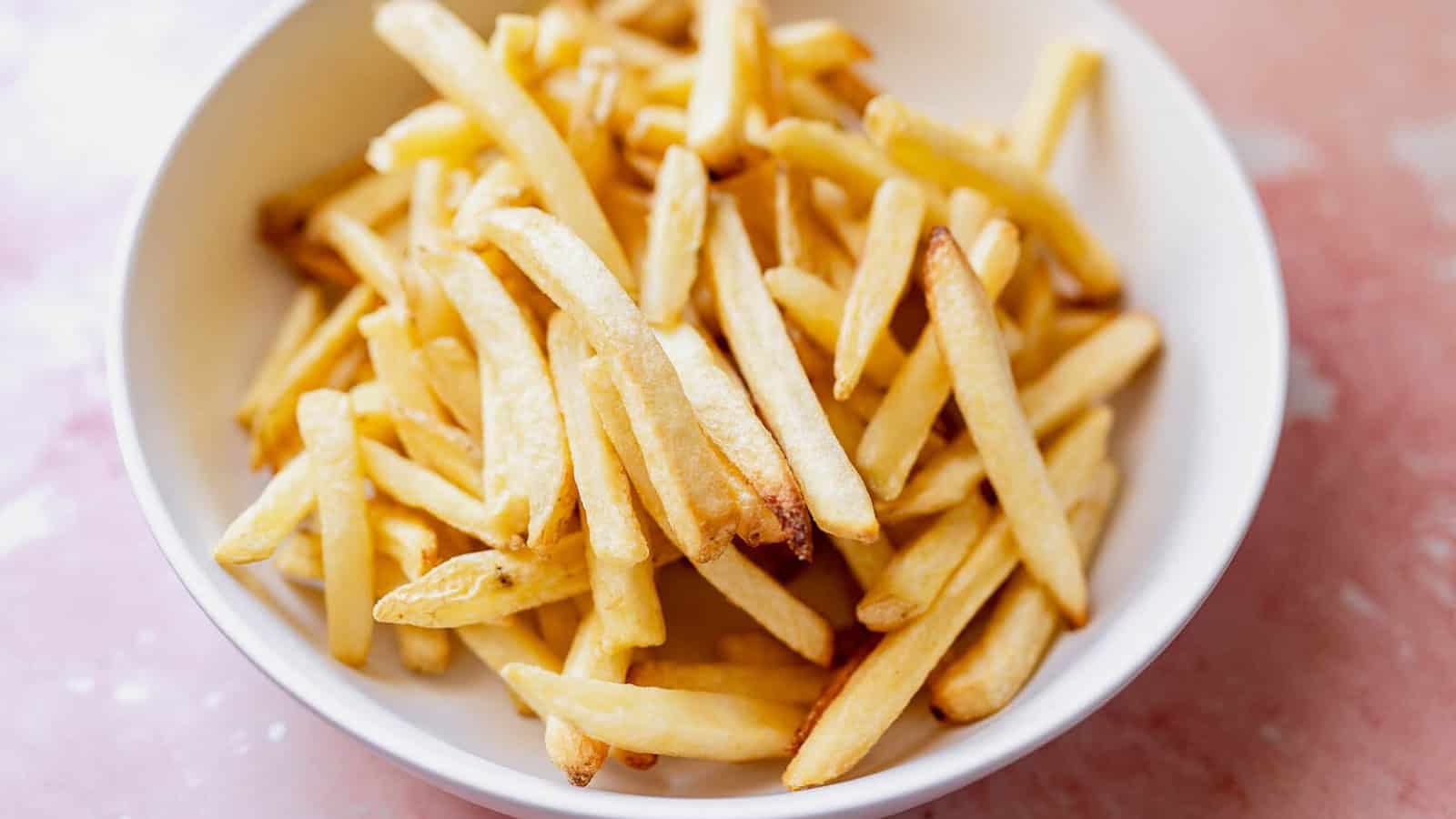 A white bowl filled with golden-brown French fries on a pinkish surface.