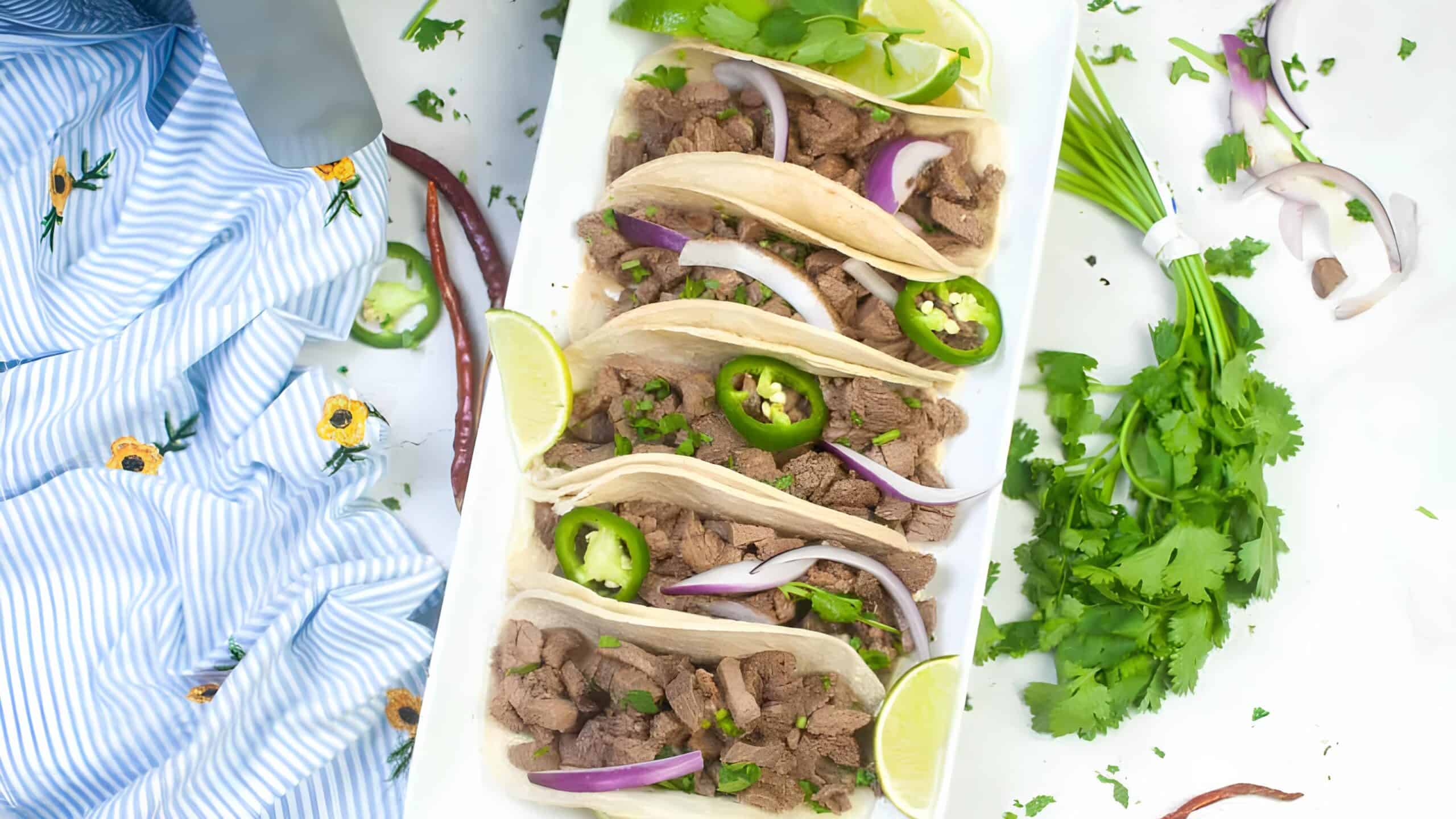 A plate of beef tacos with sliced jalapeños, red onion, and lime wedges. Fresh cilantro and a striped cloth are nearby.