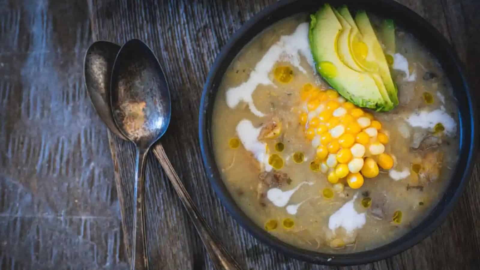 A bowl of soup topped with avocado slices and corn on the cob, alongside two spoons on a wooden surface.