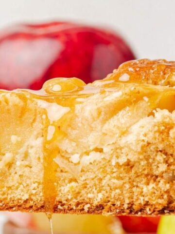 Close-up of a slice of apple cake with caramel drizzle, held up on a fork, with red apples in the background.