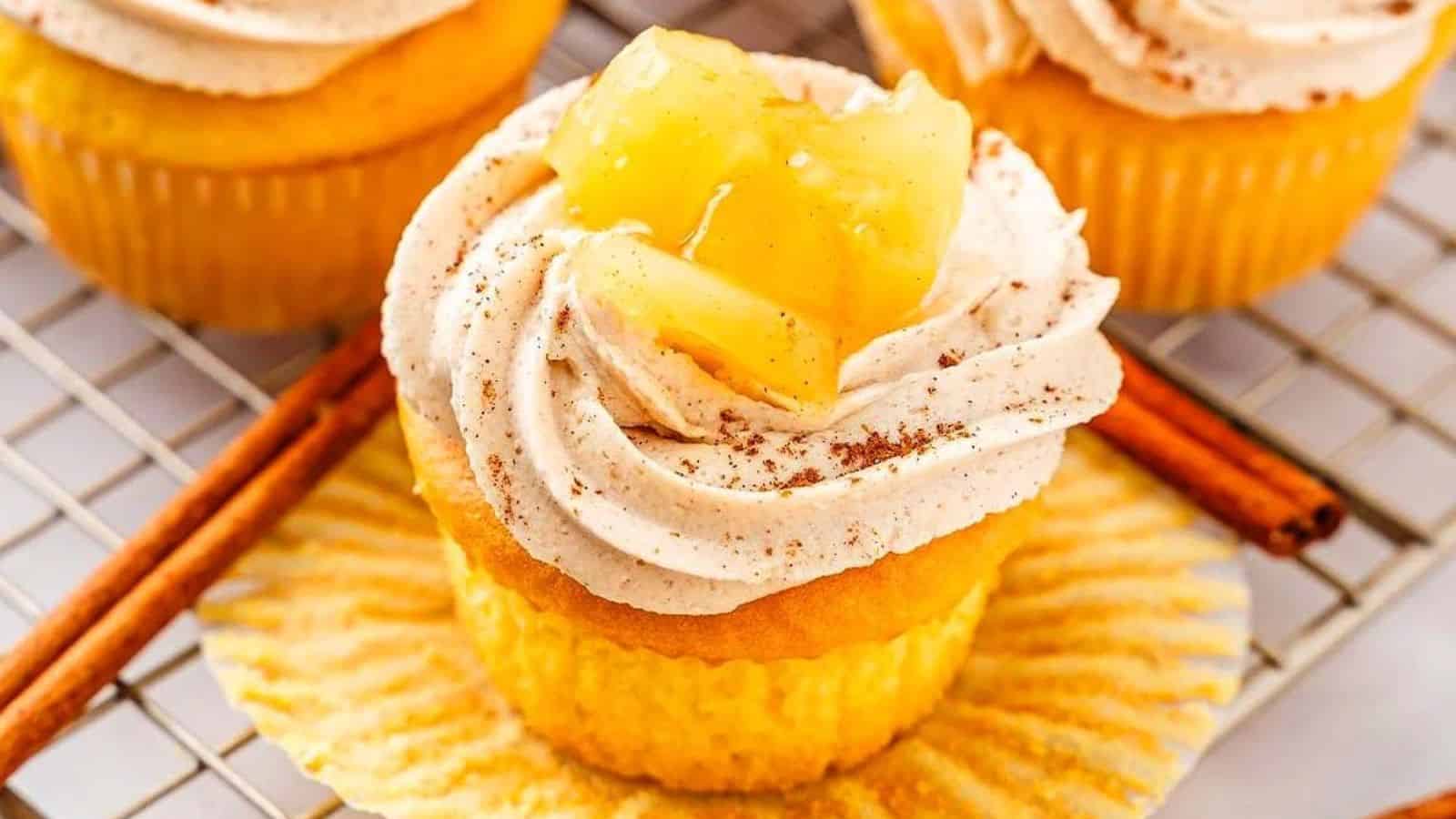 Cupcake with cream topping and pineapple pieces, placed on an open wrapper, with cinnamon sticks in the background.