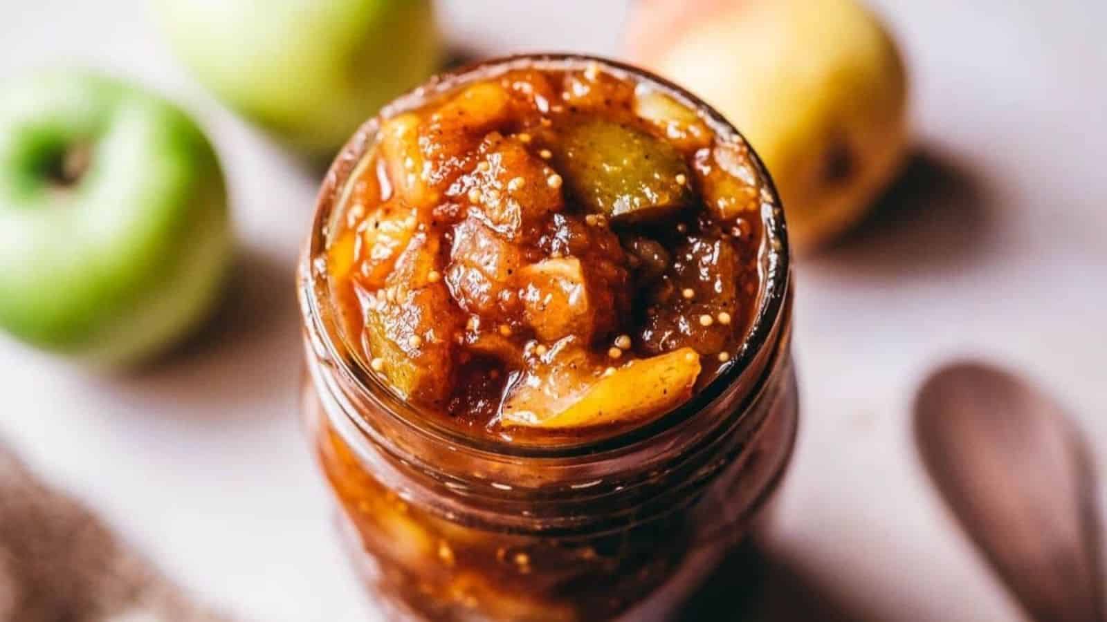 A jar of chunky apple chutney with visible pieces of apple, surrounded by a few whole green apples.